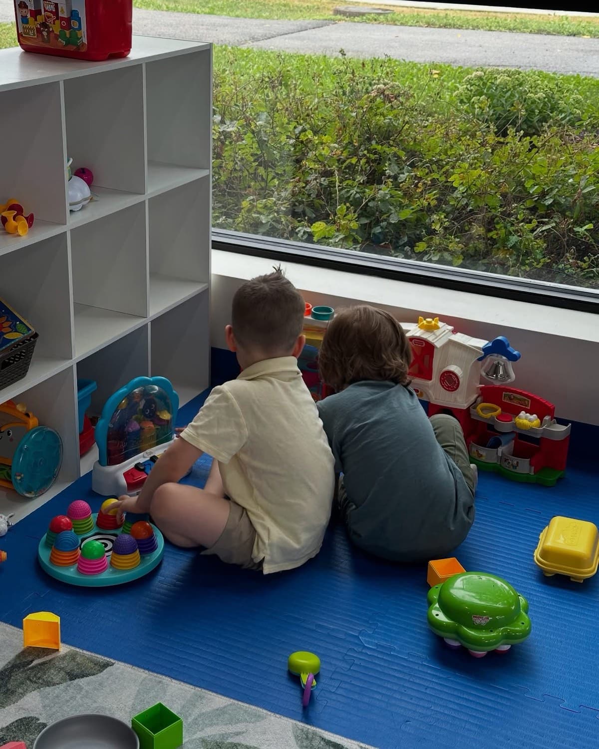 Two children sitting side by side on a playroom floor, looking out a window together.