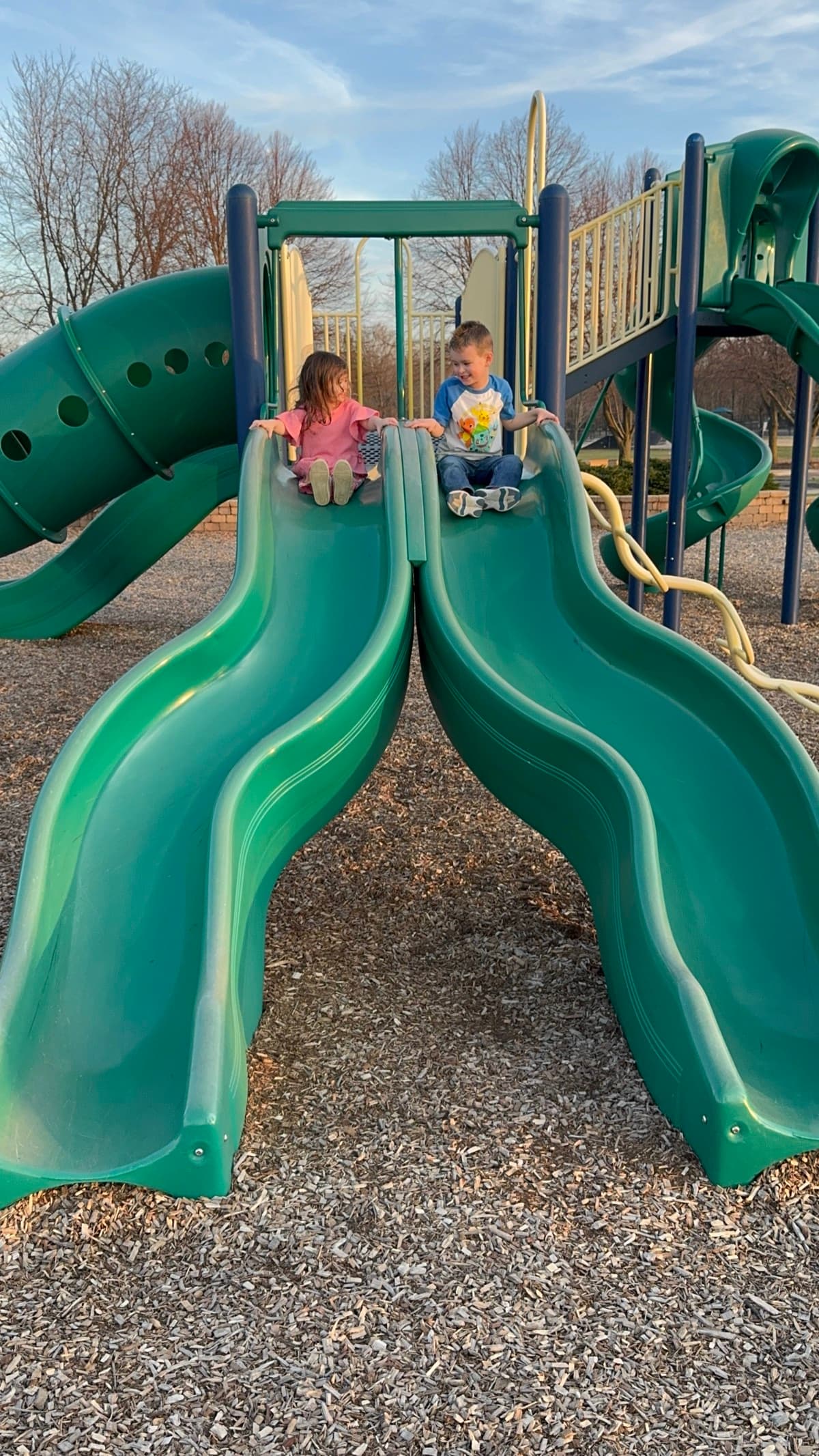 Two children sitting at the top of side-by-side playground slides, smiling at each other.